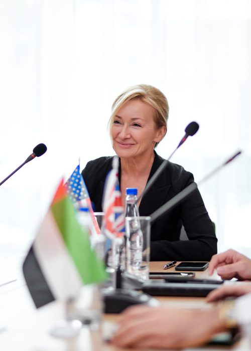 female delegate in formalwear looking at audience, giving speech report at multi-ethnic conference, meeting in boardroom, using microphones